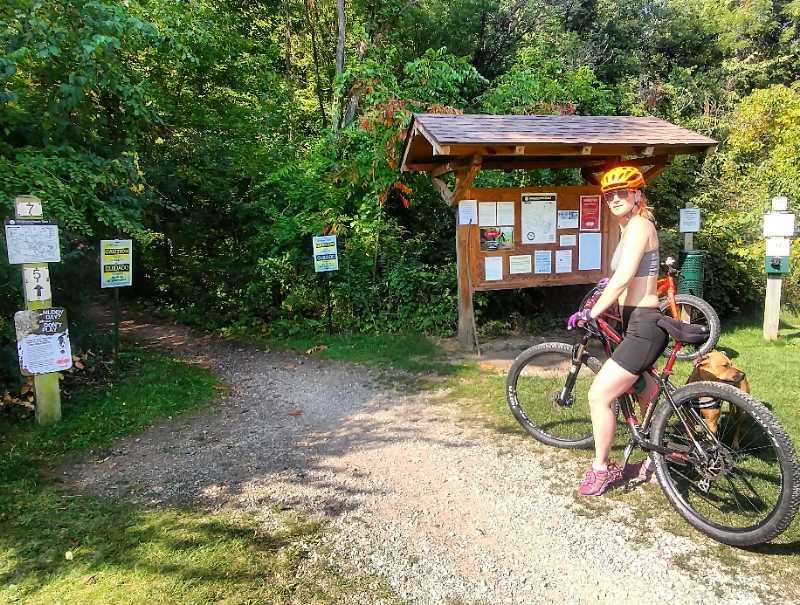 A person wearing a helmet and athletic attire stands beside a mountain bike at a trailhead. A wooden information signboard provides details about the trails, while surrounding greenery showcases a lush, natural environment. A dog is sitting nearby. Potawatomi trail mountain bike trail.