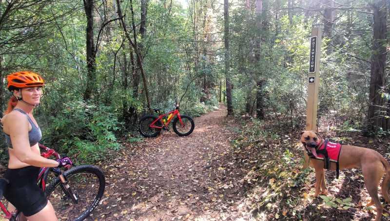 A woman wearing a helmet and athletic clothing stands beside a mountain bike on a dirt trail in a wooded area. Nearby, a brown dog with a red harness is sitting, and a trail sign points towards different paths. The scene is surrounded by lush green foliage and autumn leaves scattered on the ground. Potawatomi trail mountain bike trail.