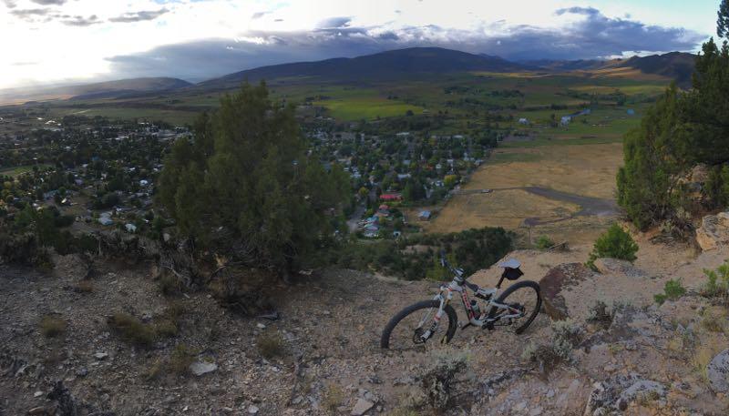 A scenic view from a rocky hillside overlooking a small town and lush green fields, with a mountain bike resting on the ground in the foreground. The sky features scattered clouds and a hint of sunset light, creating a peaceful outdoor atmosphere. Dorcas-Jensen Park mountain bike trail.