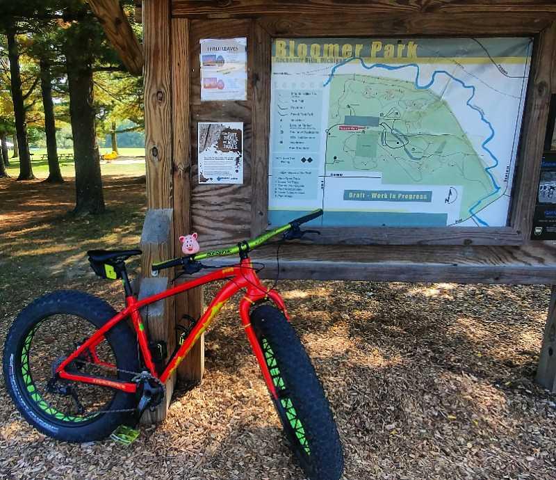 A vibrant red and green fat bike is leaning against a wooden park sign, which features a map of Bloomer Park. The map outlines the trails available and includes various informational flyers attached to the sign. In the background, lush trees and a grassy area can be seen, creating a scenic outdoor environment. Bloomer Park mountain bike trail.