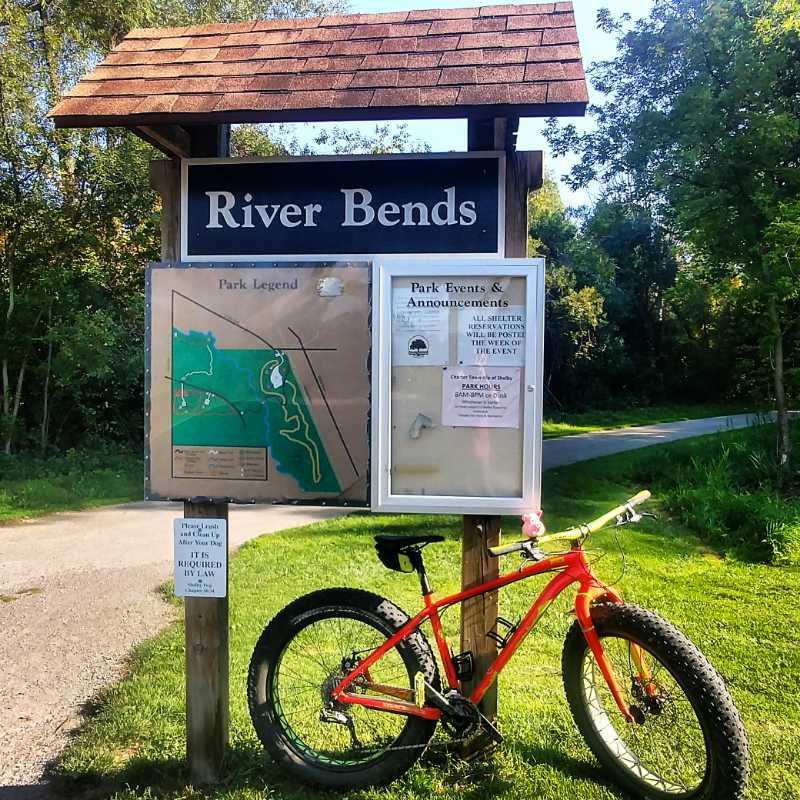 A sign for River Bends park with a map legend and announcements board, accompanied by an orange fat tire bike parked on the grass. The background features trees and a winding path. River Bends Mountain Bike Trail mountain bike trail.