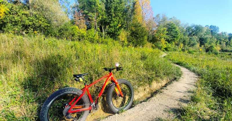 A vibrant red fat bike rests beside a winding dirt path, surrounded by tall green grass and trees with autumn foliage. The scene is bright and sunny, showcasing a peaceful outdoor environment perfect for biking adventures. River Bends Mountain Bike Trail mountain bike trail.
