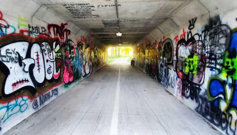 A vibrant urban tunnel adorned with colorful graffiti on the walls, leading towards a brightly lit exit. The floor features a white line, suggesting a path for pedestrians or cyclists. The scene is filled with various artistic expressions and tags, creating an energetic atmosphere. River Bends Mountain Bike Trail mountain bike trail.