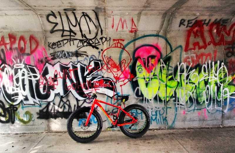 A vibrant scene featuring a bright red bike positioned in front of a colorful graffiti-covered wall. The wall displays various tags and designs in shades of blue, pink, green, and black, creating an urban, artistic backdrop. The overall atmosphere conveys a sense of creativity and street culture. River Bends Mountain Bike Trail mountain bike trail.
