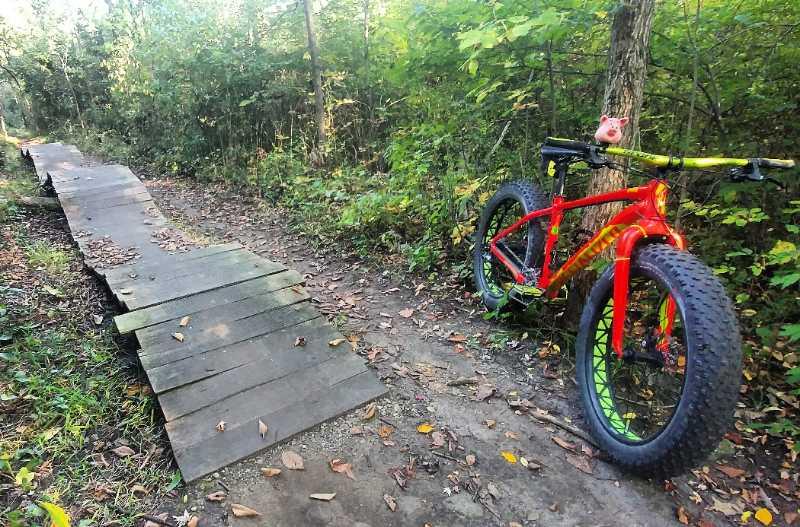 A vibrant red mountain bike with thick tires is parked next to a wooden plank trail in a lush green forest. The trail, lined with fallen leaves, leads into the woods, inviting exploration. Sunlight filters through the trees, enhancing the serene natural setting. River Bends Mountain Bike Trail mountain bike trail.