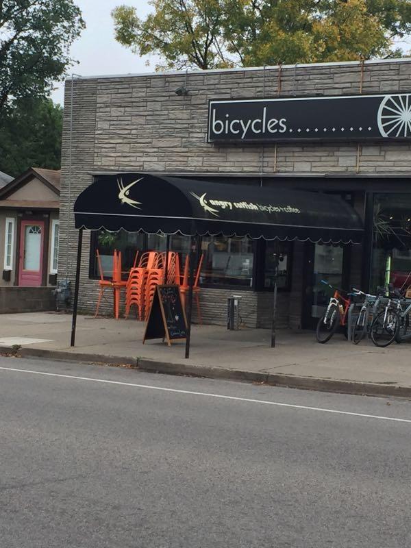 A street view of a bicycle shop with a black awning featuring the name "angry manita..." in gold text. There are orange stacked chairs beneath the awning and a sign nearby. In front of the shop, several bicycles are displayed. A residential building with a pink door is visible to the left. Trees and a cloudy sky are in the background.