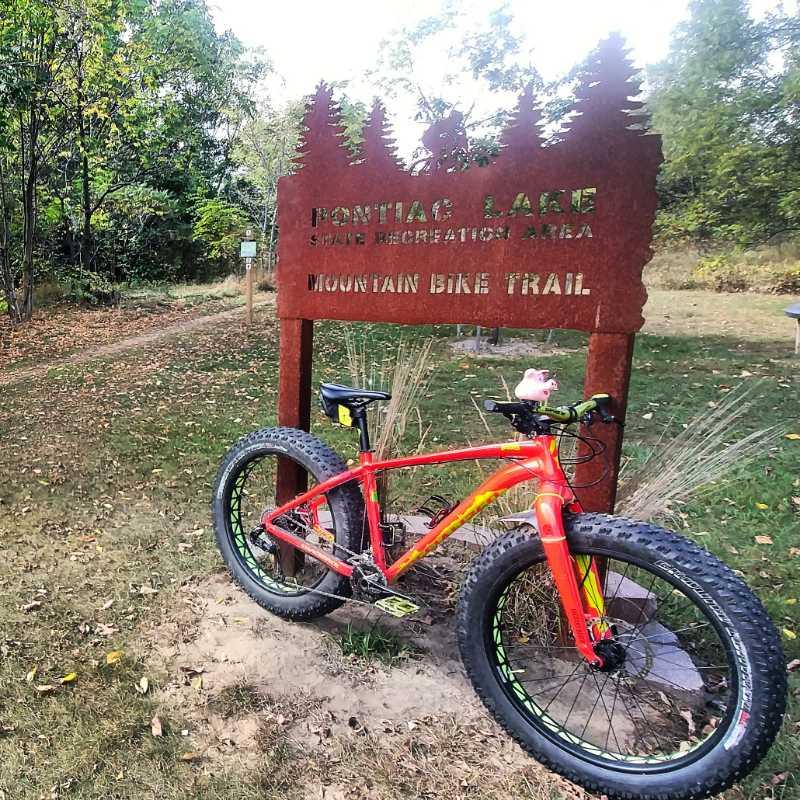 A brightly colored mountain bike leaning against a rustic sign that reads "Pontiac Lake State Recreation Area Mountain Bike Trail." The background features a natural setting with trees and grassy areas, indicating a scenic outdoor environment. Pontiac Lake mountain bike trail.