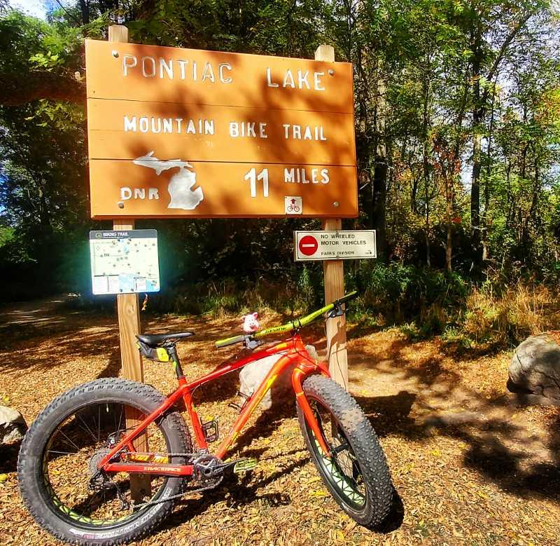 A sign at the entrance of the Pontiac Lake Mountain Bike Trail, indicating an 11-mile route. A bright orange mountain bike is leaning against the sign, surrounded by trees and fallen leaves. Additional signage suggests no wheeled motor vehicles are allowed on the trail. Pontiac Lake mountain bike trail.