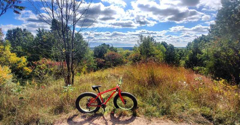 A bright red bike rests on a dirt path surrounded by tall grass and colorful foliage. In the background, trees and a wide, open sky with fluffy clouds can be seen. The scene captures the beauty of nature on a sunny day. Pontiac Lake mountain bike trail.