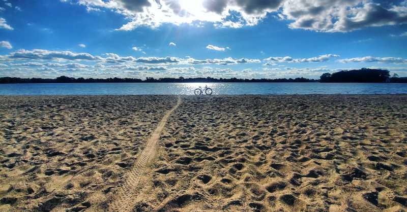 A sandy beach with a calm body of water in the background, under a bright blue sky filled with fluffy clouds. A bicycle is parked near the water's edge, and a pathway of footprints leads through the sand towards the water. Sunlight glistens on the surface of the water, creating a serene and peaceful atmosphere. Pontiac Lake mountain bike trail.