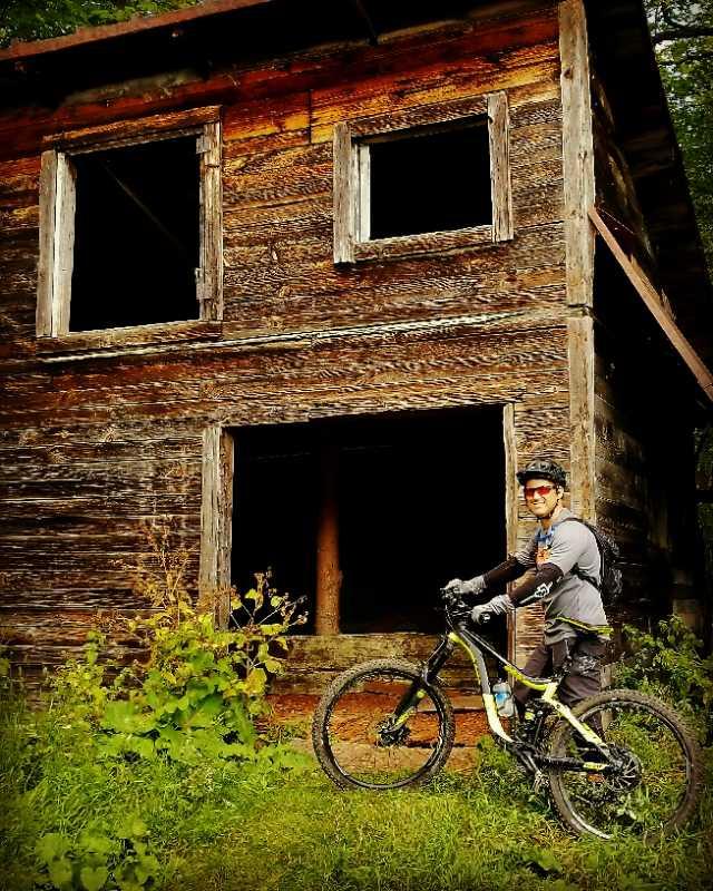 A mountain biker wearing a helmet and sunglasses stands next to a bicycle in front of a rustic, weathered wooden cabin with empty windows, surrounded by greenery and tall grass. Sentiers de l'Abbaye d'Oka mountain bike trail.