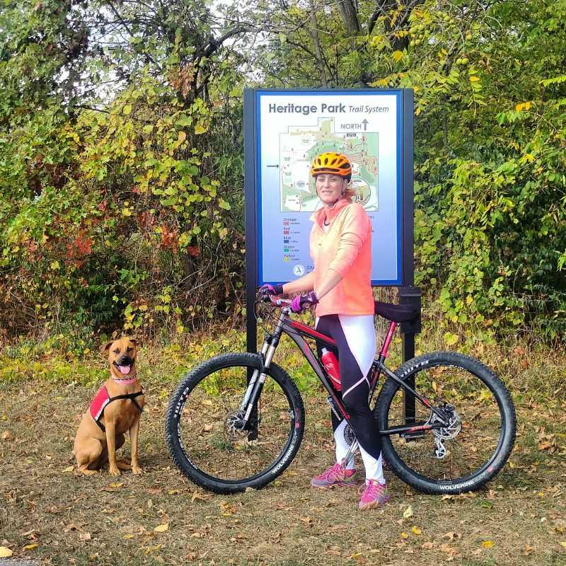 A woman in a bright orange long-sleeve shirt and a helmet stands next to her mountain bike, smiling at the camera. Beside her, a brown dog wearing a red harness sits happily. In the background, there is a sign displaying a map of the Heritage Park Trail System, surrounded by trees with autumn foliage. The setting conveys a fun outdoor adventure spirit. Heritage Park mountain bike trail.