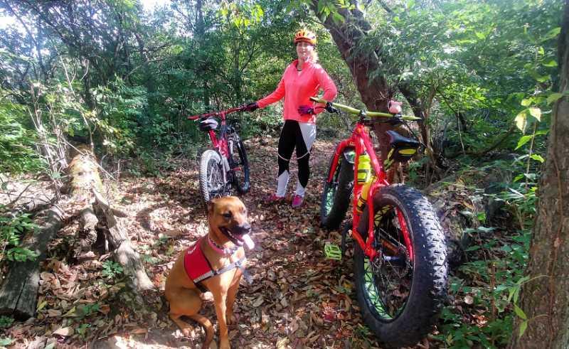 A person in a bright pink jacket and a helmet stands between two bicycles in a wooded area filled with greenery. One bicycle is red and the other is black, with large tires. A brown dog with a harness sits calmly in front of the bikes, surrounded by fallen leaves on the forest floor. Heritage Park mountain bike trail.