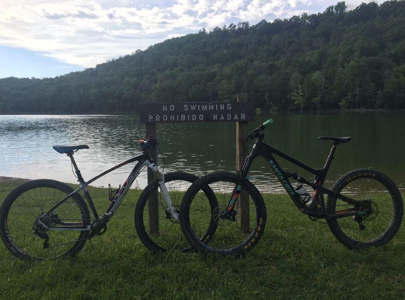 Two mountain bikes parked on a grassy area near a lake. In the background, a sign reads "No Swimming" in English and Spanish ("Prohibido Nadar"). Lush green trees and hills are visible across the water under a partly cloudy sky. Panther Creek State Park mountain bike trail.