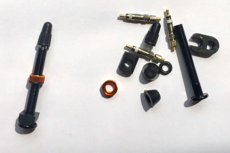 Assorted bike valve components, including a valve body with an orange ring, metal and rubber washers, black plastic caps, and other small fittings scattered on a white background.
