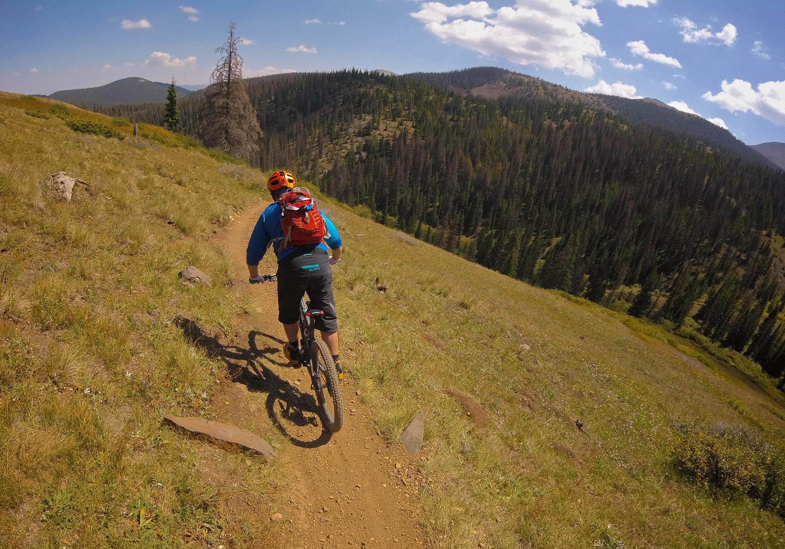 A mountain biker riding along a winding dirt trail through a green, grassy landscape with trees and hills in the background. The cyclist is wearing a blue jacket, black shorts, and an orange helmet, with a red backpack on their back. The sky is bright and partly cloudy. Monarch Crest Trail mountain bike trail.