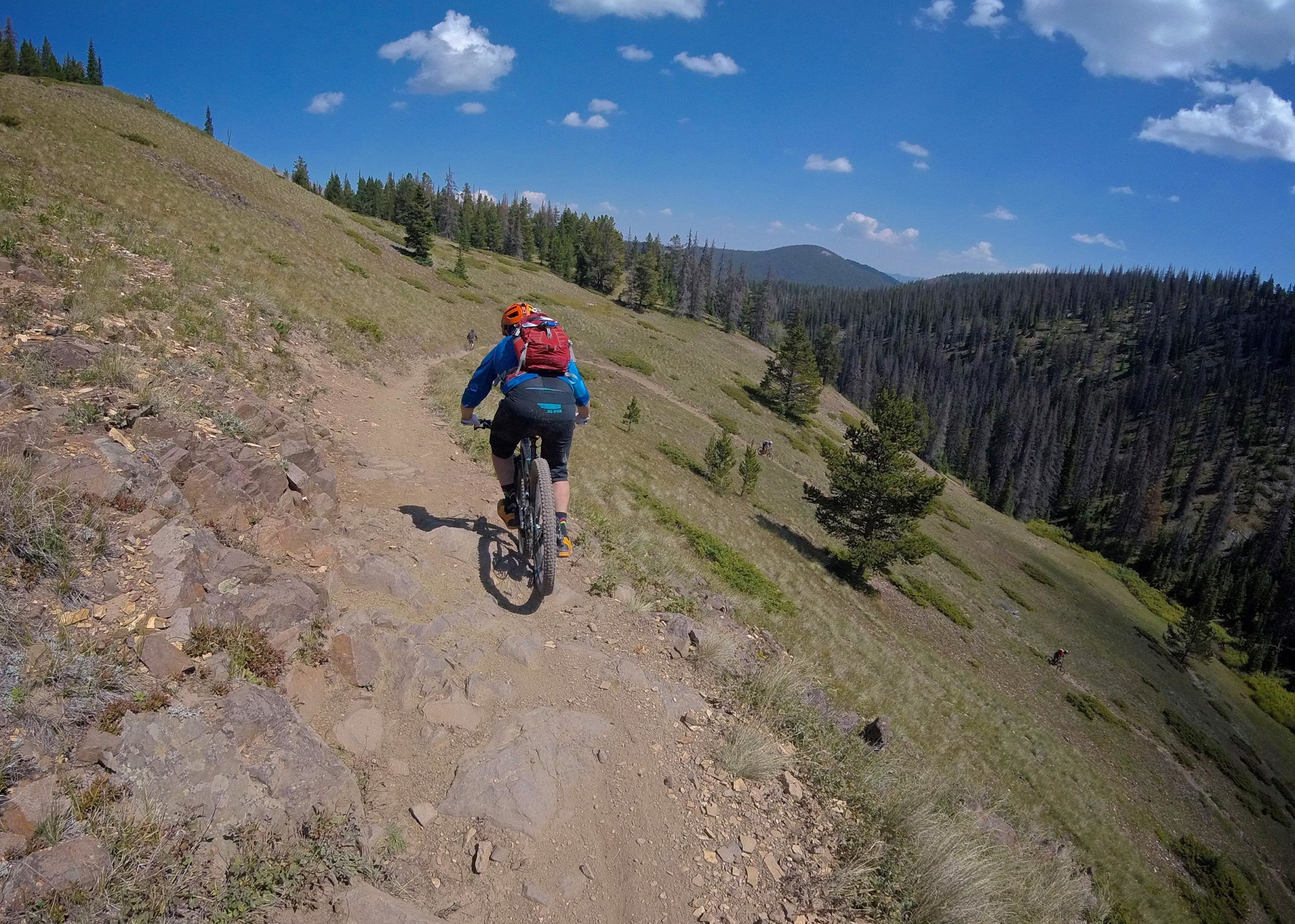 A person riding a mountain bike on a rocky trail through a green landscape, surrounded by trees and under a blue sky with scattered clouds. Monarch Crest Trail mountain bike trail.