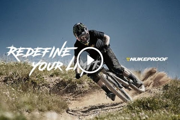 A mountain biker skillfully navigating a dirt trail on a sunny day, surrounded by greenery. The image features dynamic movement and dust being kicked up, with the text "REDEFINE YOUR LIMITS" and the Nukeproof logo overlaid.