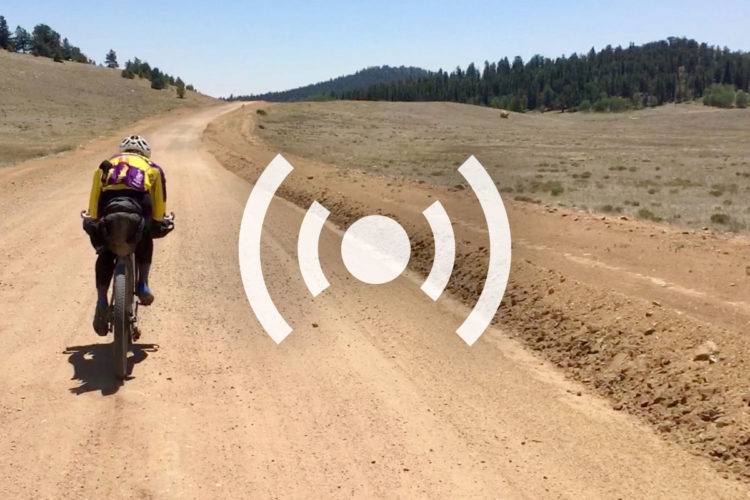 A cyclist in a yellow and purple jersey rides a bike along a dirt road surrounded by open fields and low hills. Clear blue skies and distant trees can be seen in the background. A semi-transparent graphic indicating a sound signal is overlaid in the center of the image.