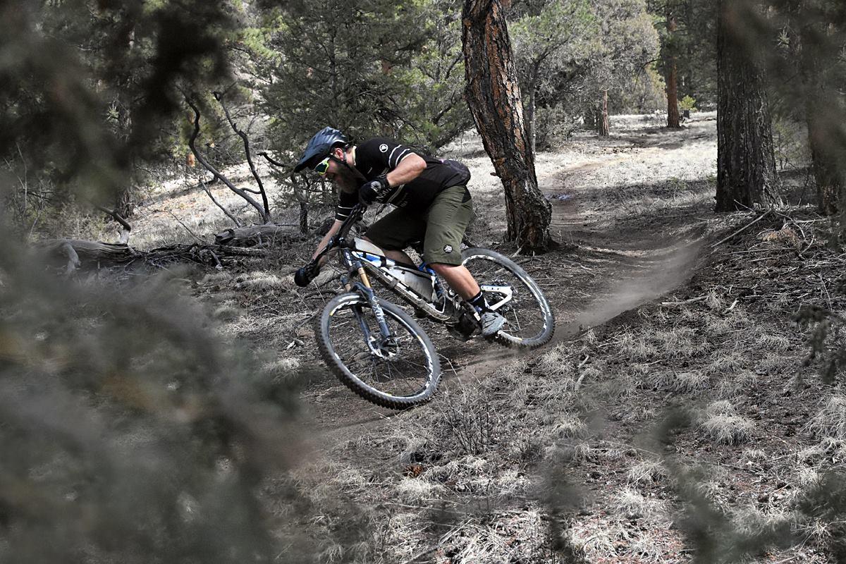 Michelin Wild AM: A mountain biker skillfully navigates a dirt trail through a forest, leaning into a turn while kicking up dust. Pine trees and brush frame the scene, capturing the excitement of outdoor cycling.