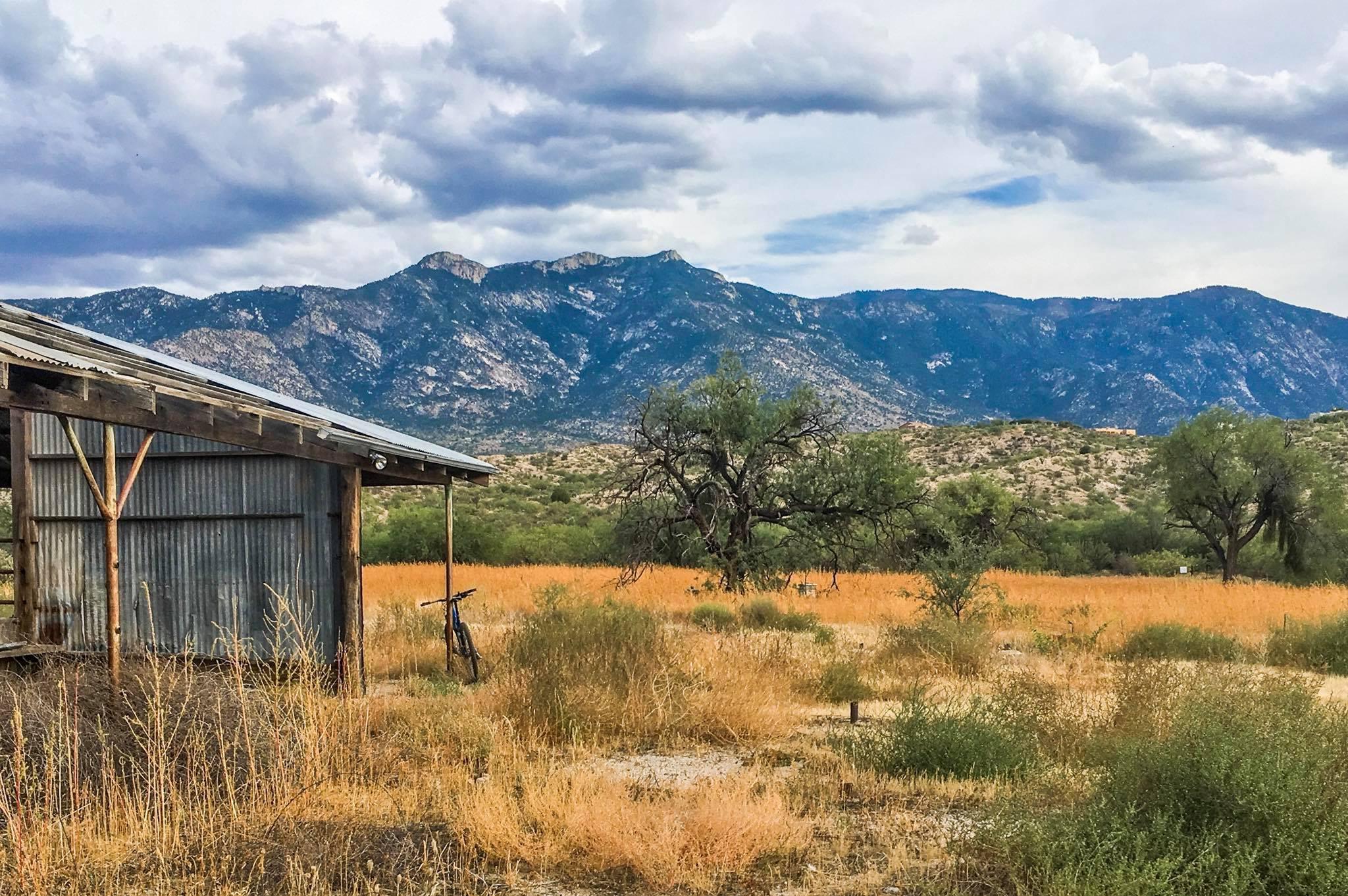A rustic barn made of corrugated metal stands in a dry, grassy field, with a bicycle leaning against it. In the background, towering mountains rise under a partly cloudy sky, showcasing a natural landscape of rolling hills and sparse vegetation. 50-year Trail / Golder Ranch mountain bike trail.