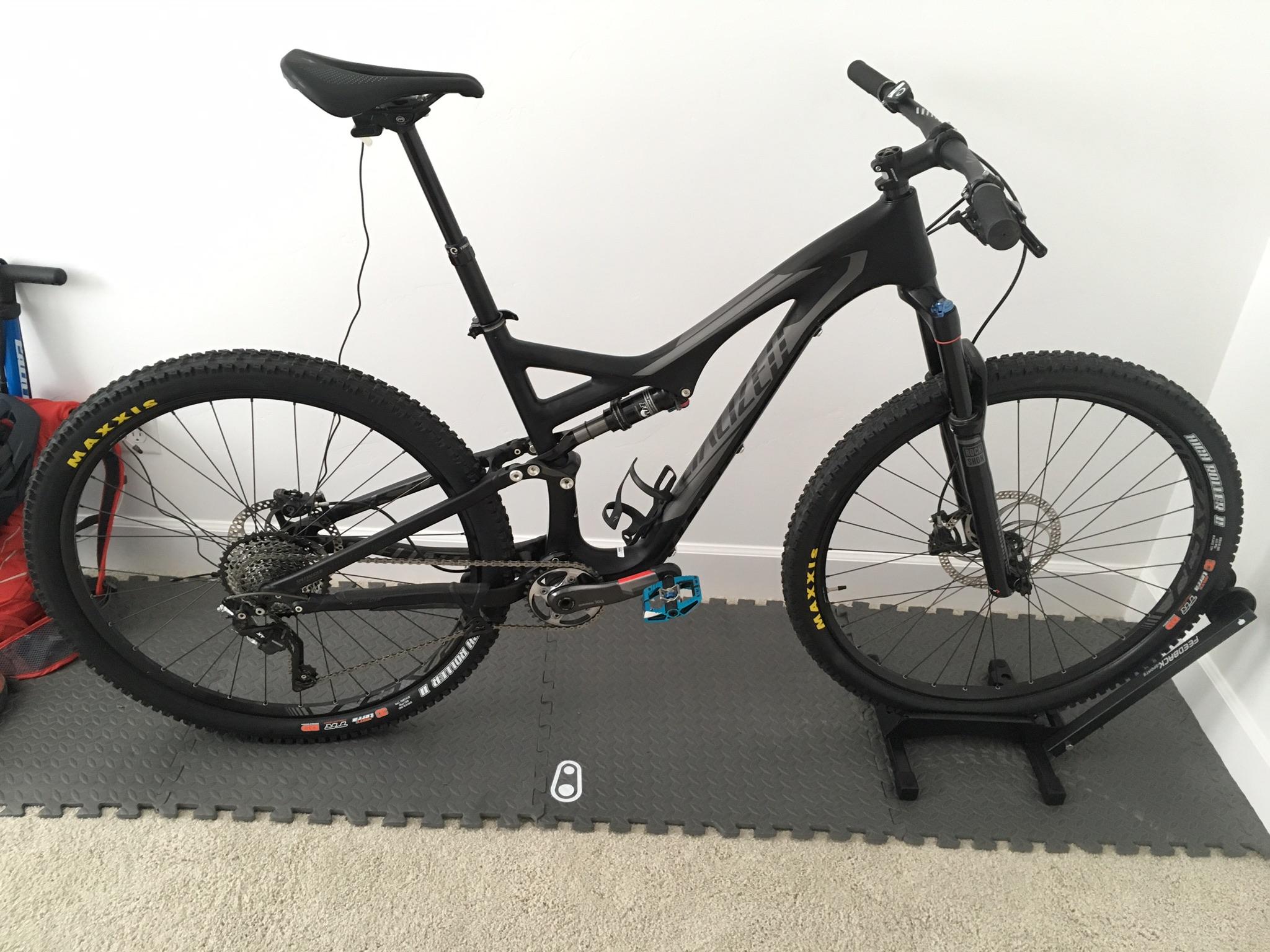 Specialized Stumpjumper FSR comp carbon 650B: A black mountain bike with a sleek frame design is displayed on a bike stand. The bike features thick tires with yellow "Maxxis" branding, a dropper seat post, and a modern gear system. It is positioned against a white wall, with a rubber mat underneath and a glimpse of a red backpack in the background.