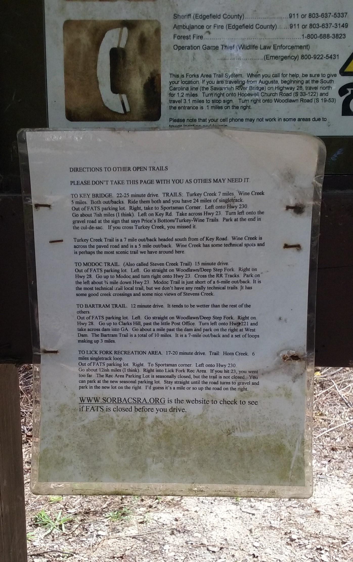 A weathered sign providing directions to various open trails in the Forks Area Trail System. The sign includes specific driving instructions and distances for reaching multiple trails, such as Turkey Creek, Modoc Trail, Bartram Trail, and Lick Fork Recreation Area, along with a note advising not to take the page. Additionally, it features emergency contact numbers and a website link for trail closures. The text is partially faded and the sign is mounted on a wooden post in a natural setting. Forks Area Trail System (FATS) mountain bike trail.