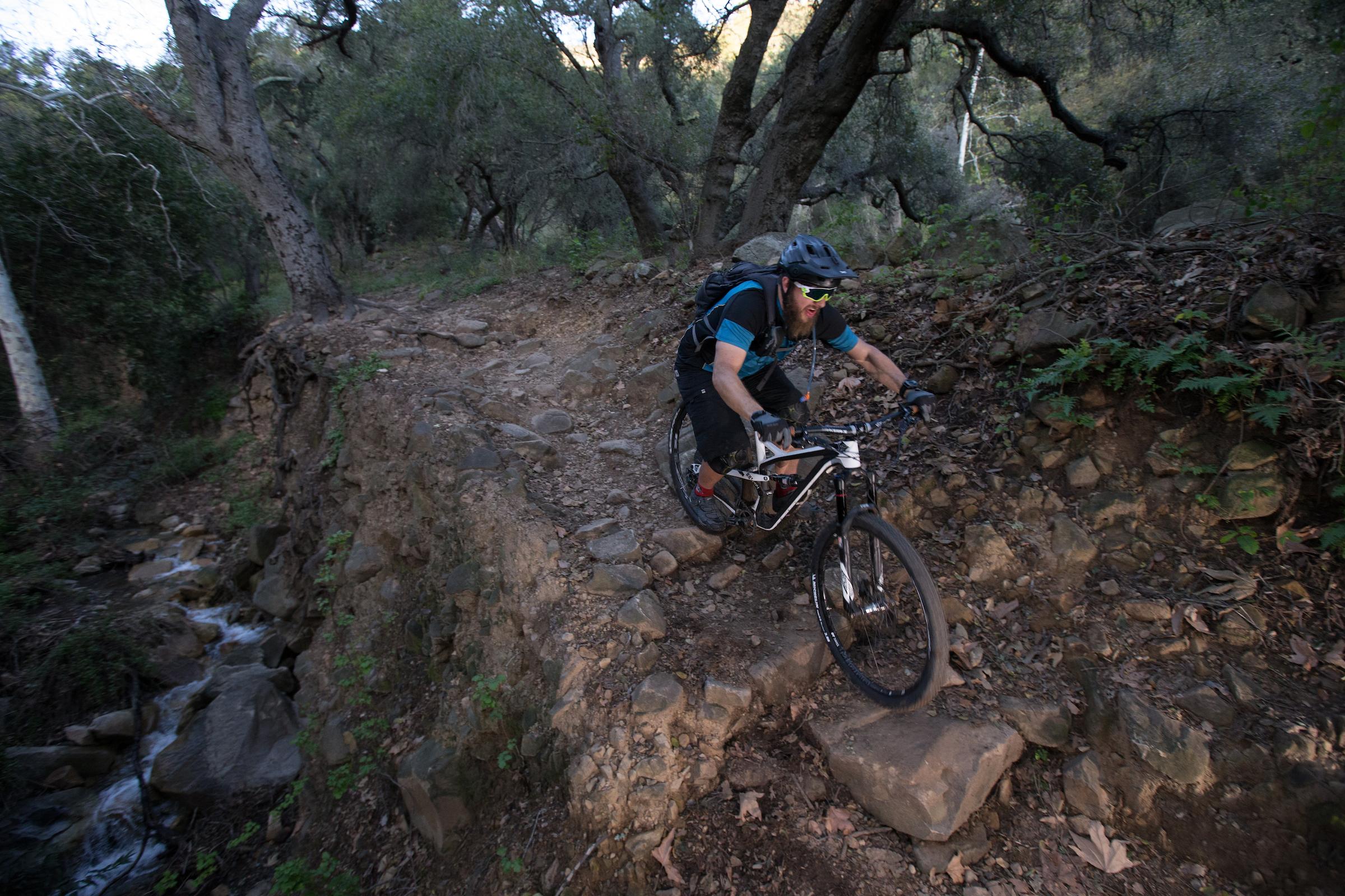 Michelin Wild AM: A mountain biker navigating a rocky trail in a forested area. The rider is wearing a helmet and sunglasses, focused on maintaining balance as he approaches a steep section of the path. Surrounding him are trees and greenery, with a small creek visible to the left.