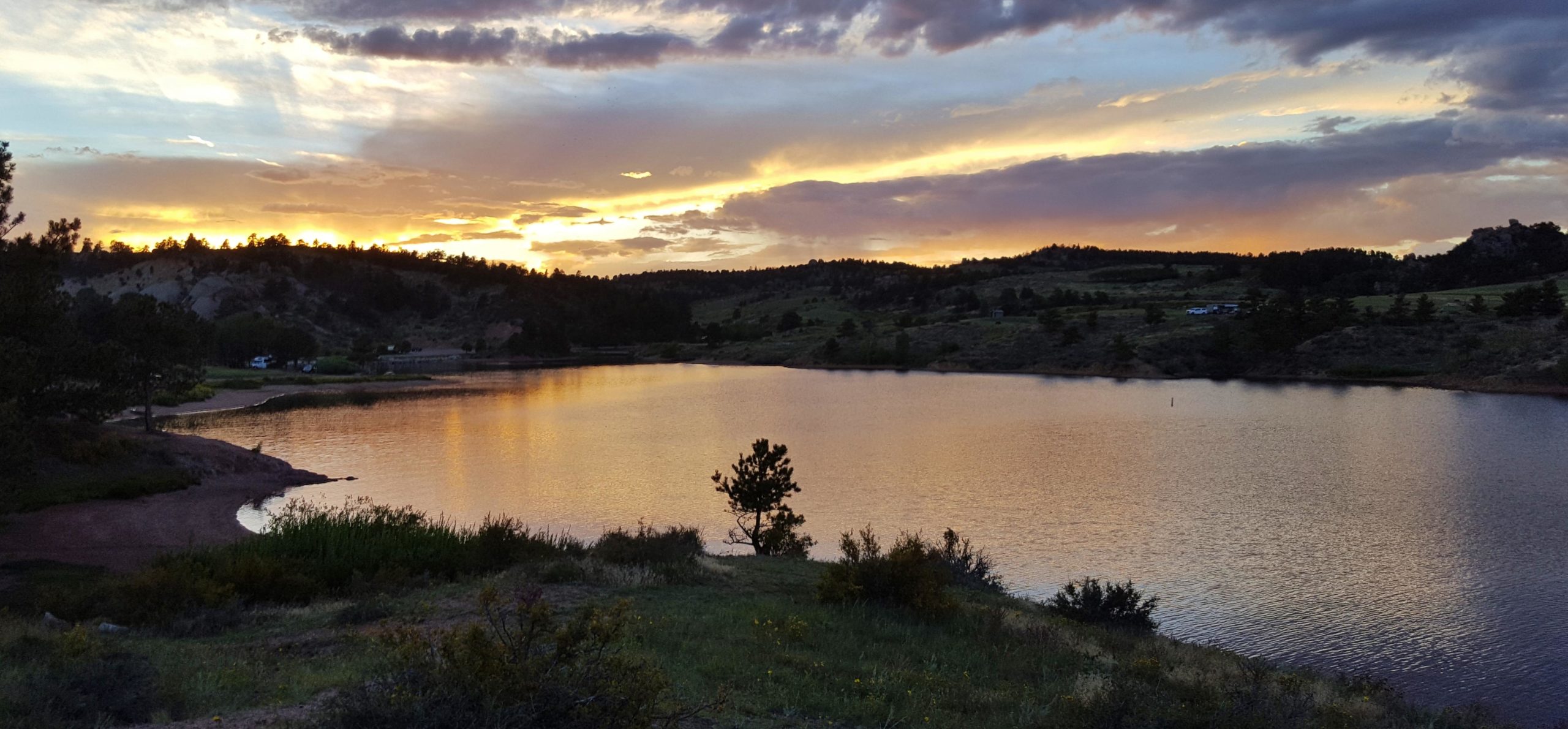 A scenic sunset view over a calm lake, surrounded by lush greenery and gently rolling hills. The sky is adorned with shades of orange and purple, reflecting beautifully on the water's surface. Small trees and shrubs dot the shoreline, enhancing the peaceful atmosphere of the landscape. Curt Gowdy State Park mountain bike trail.