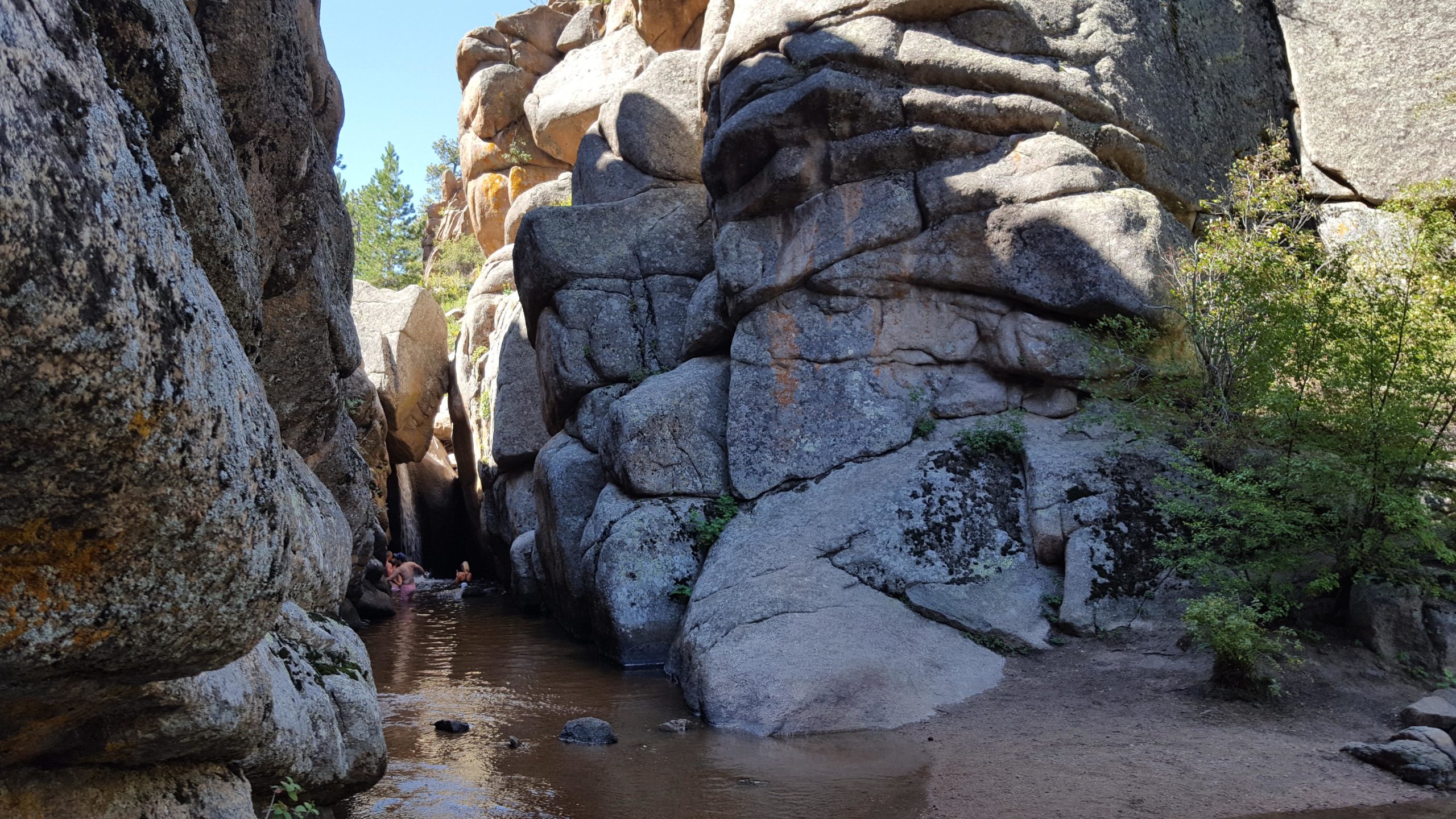 A narrow canyon with towering rock formations on either side and a shallow pool of water at the bottom. Lush greenery is visible on the right, and a few people can be seen enjoying the water. The scene is set under a clear blue sky. Curt Gowdy State Park mountain bike trail.