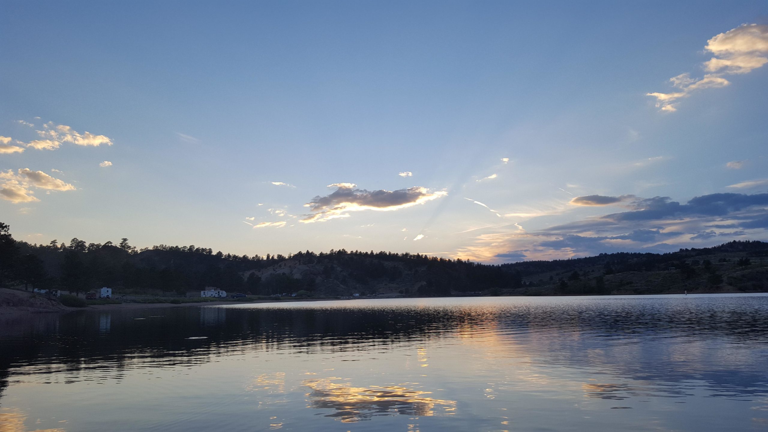A serene lake at sunset, reflecting the colorful clouds and sky. Trees line the shore, and distant hills silhouette against the fading light. The calm water captures the hues of sunset, creating a peaceful and picturesque landscape. Curt Gowdy State Park mountain bike trail.