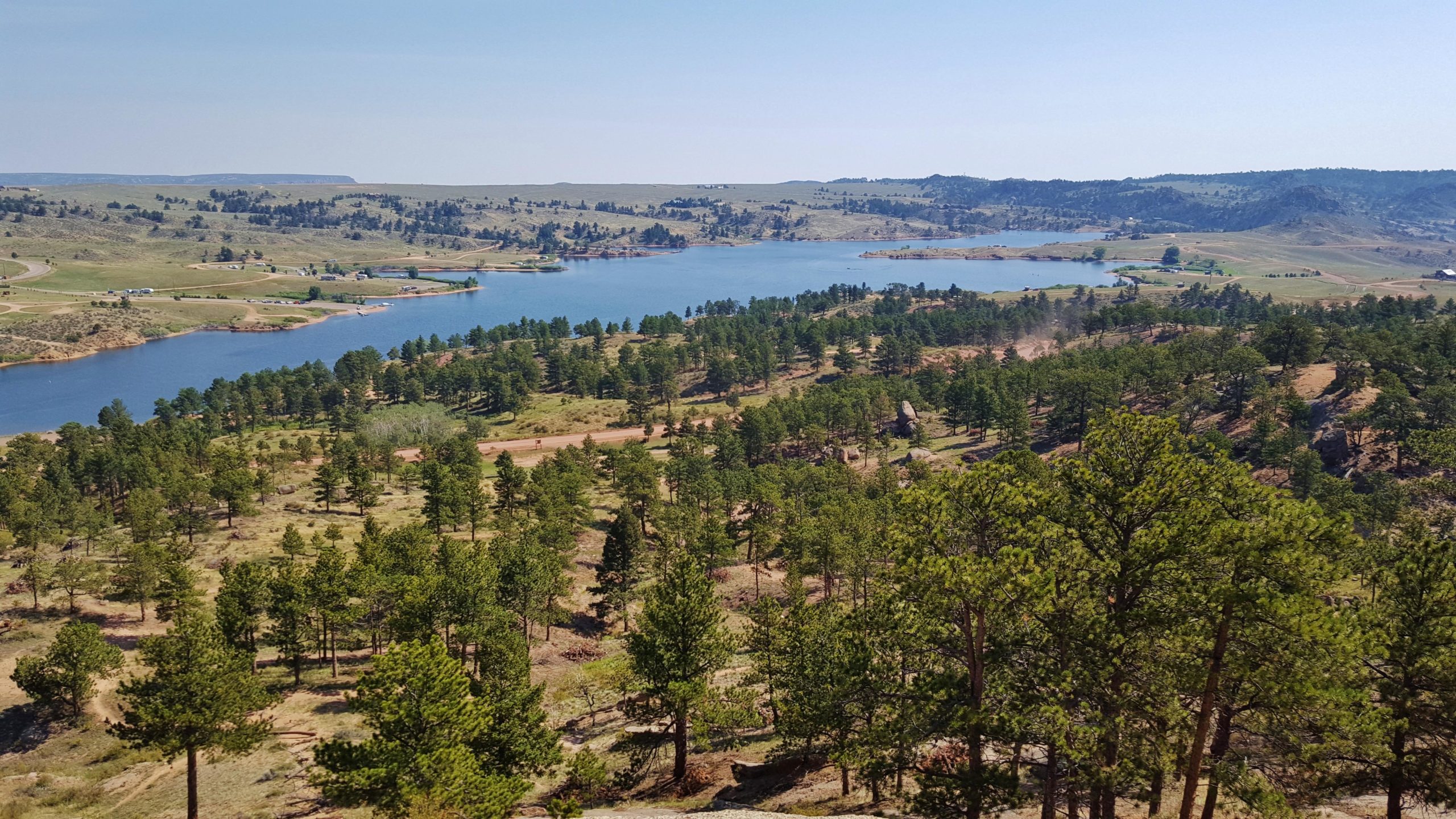 A panoramic view of a serene landscape featuring a blue lake surrounded by green pine trees, rolling hills, and distant rocky formations under a clear sky. Curt Gowdy State Park mountain bike trail.