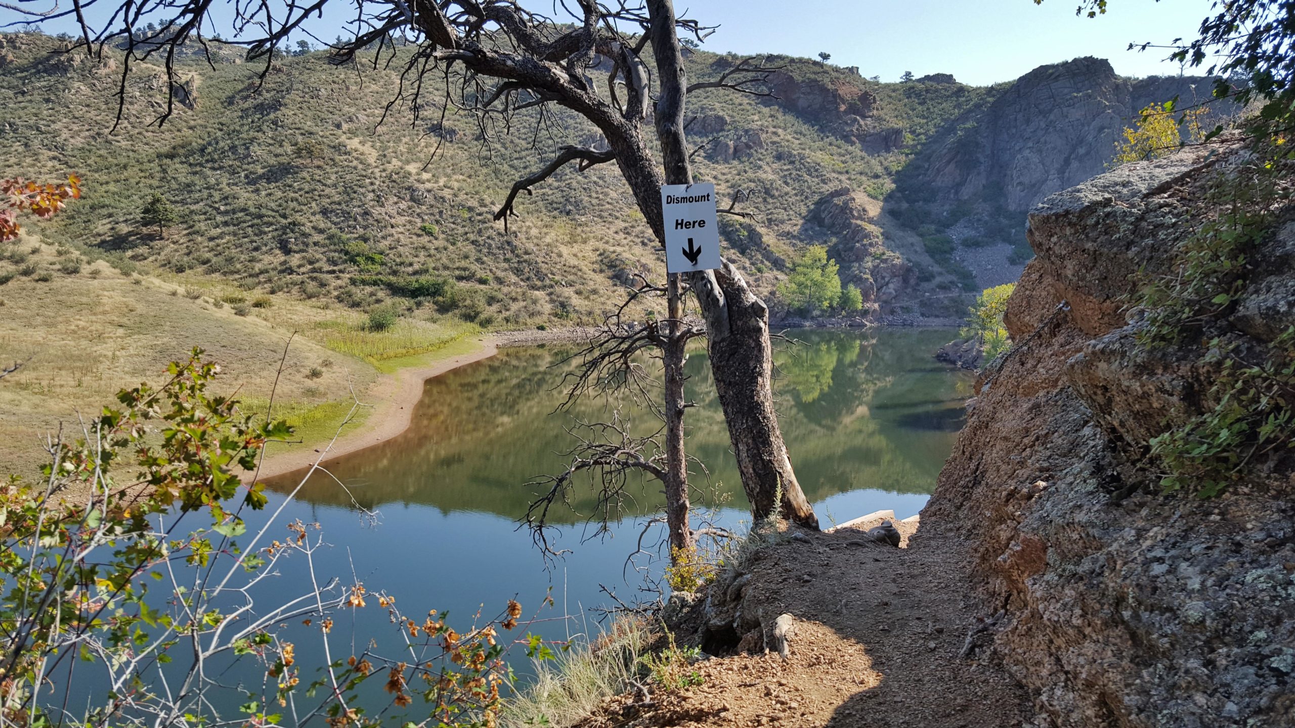 A scenic view of a tranquil lake surrounded by rolling hills and rocky terrain. In the foreground, a wooden sign with an arrow points down a pathway, indicating a spot to dismount. A tree with bare branches stands nearby, and the calm water reflects the landscape, creating a peaceful atmosphere. Cliff Hanger mountain bike trail.