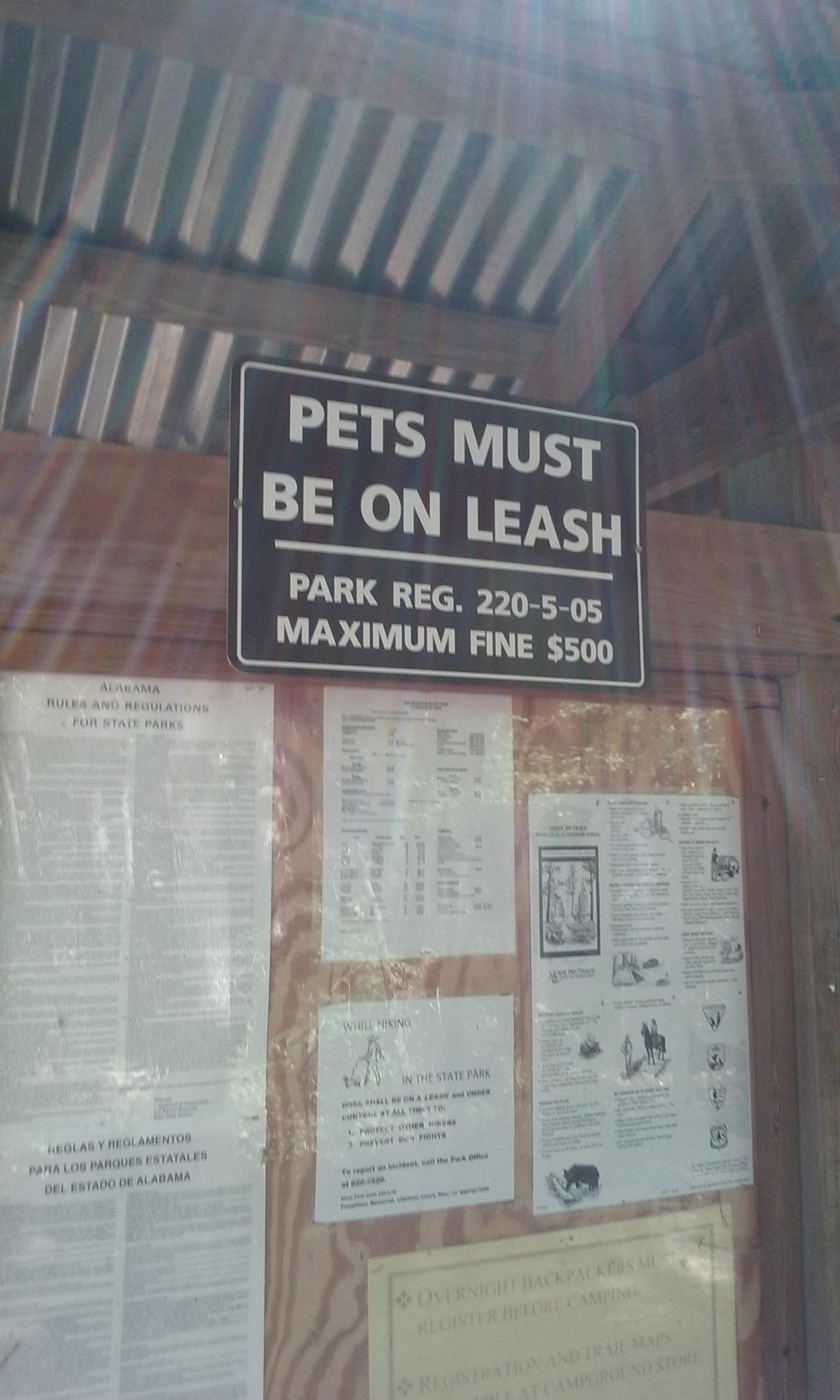 A sign displayed on a wooden structure reading "Pets Must Be On Leash" with details about park regulations, including a maximum fine of $500 for noncompliance. The background features additional informational posters about park rules and regulations. Oak Mountain State Park mountain bike trail.