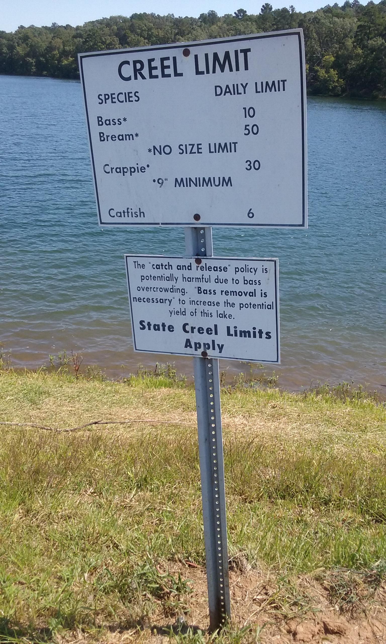 A sign displaying creel limits for fishing species, including daily limits for bass, bream, crappie, and catfish, along with information about a catch and release policy for bass. The background features a body of water and green foliage. Oak Mountain State Park Bump Trail mountain bike trail.