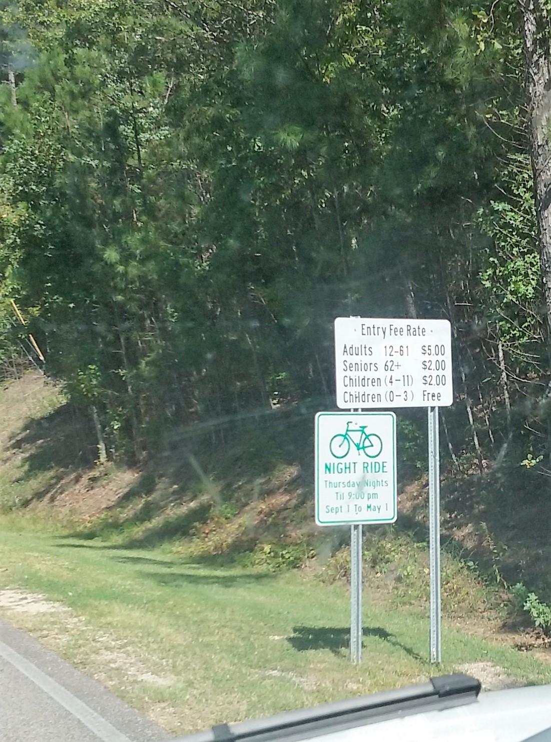A roadside sign displaying entry fee rates and information for a night ride event. The sign lists fees for adults, seniors, and children along with details about the night ride schedule. The background features a wooded area, and the sign is positioned alongside a road. Oak Mountain State Park mountain bike trail.