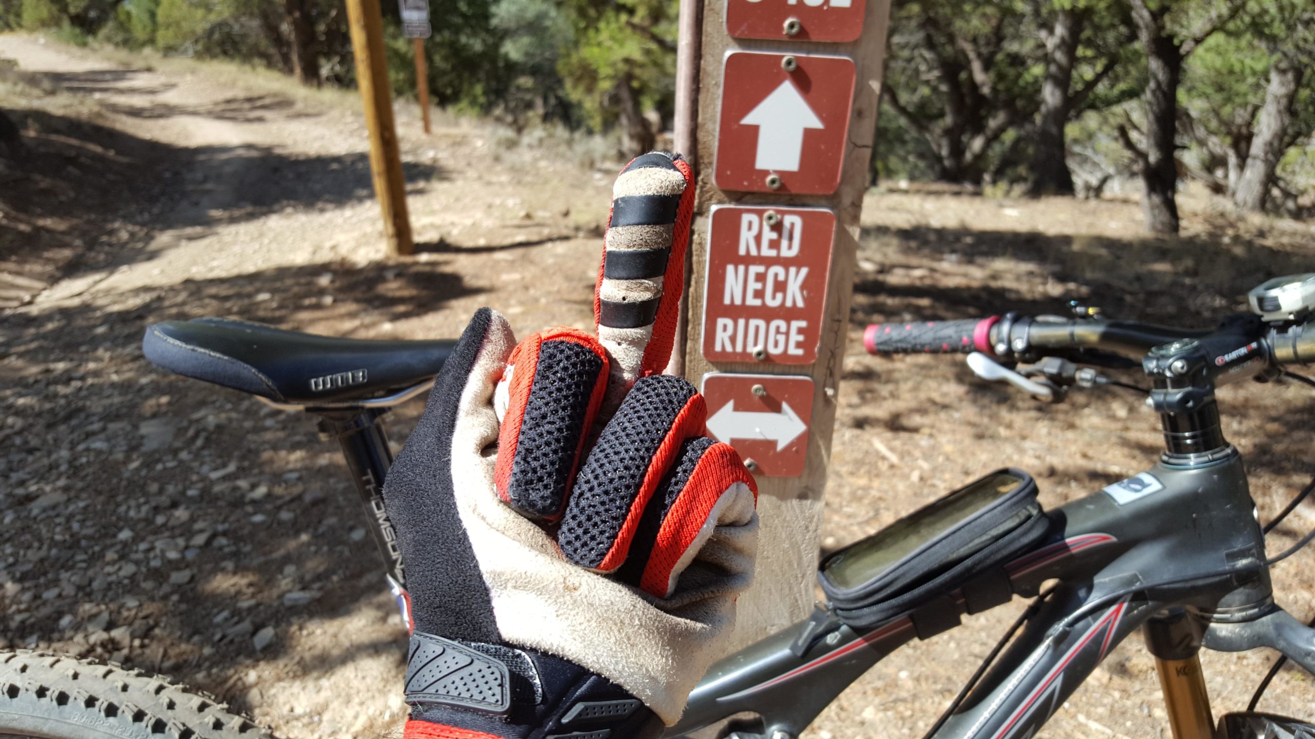A close-up of a cyclist's hand wearing black and red gloves, giving a finger gesture in front of a wooden trail sign that points to "Red Neck Ridge." A mountain bike is partially visible in the foreground, with a dirt trail and trees in the background. Redneck Ridge mountain bike trail.