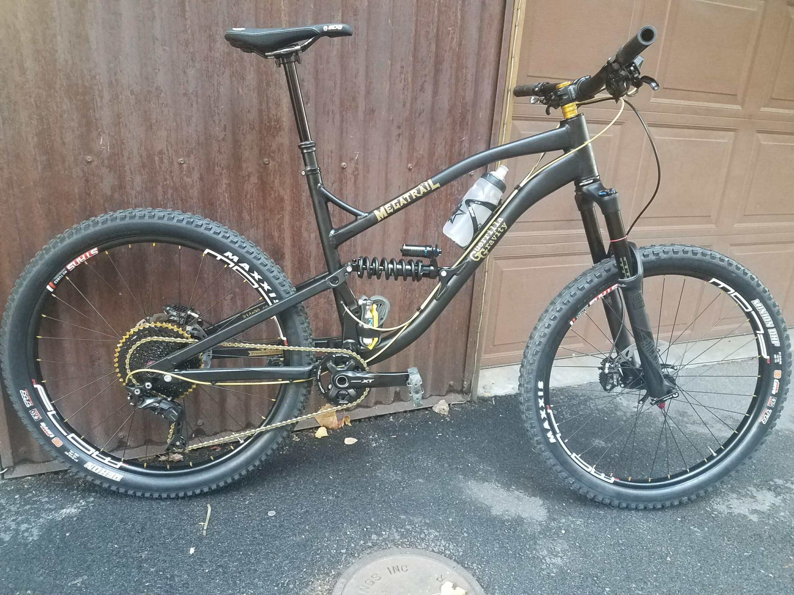 Guerrilla Gravity Megatrail: A black mountain bike with gold accents, featuring large tires, a dual suspension system, and a water bottle holder. The bike is parked against a weathered brown garage door, highlighting its sporty design and rugged components.