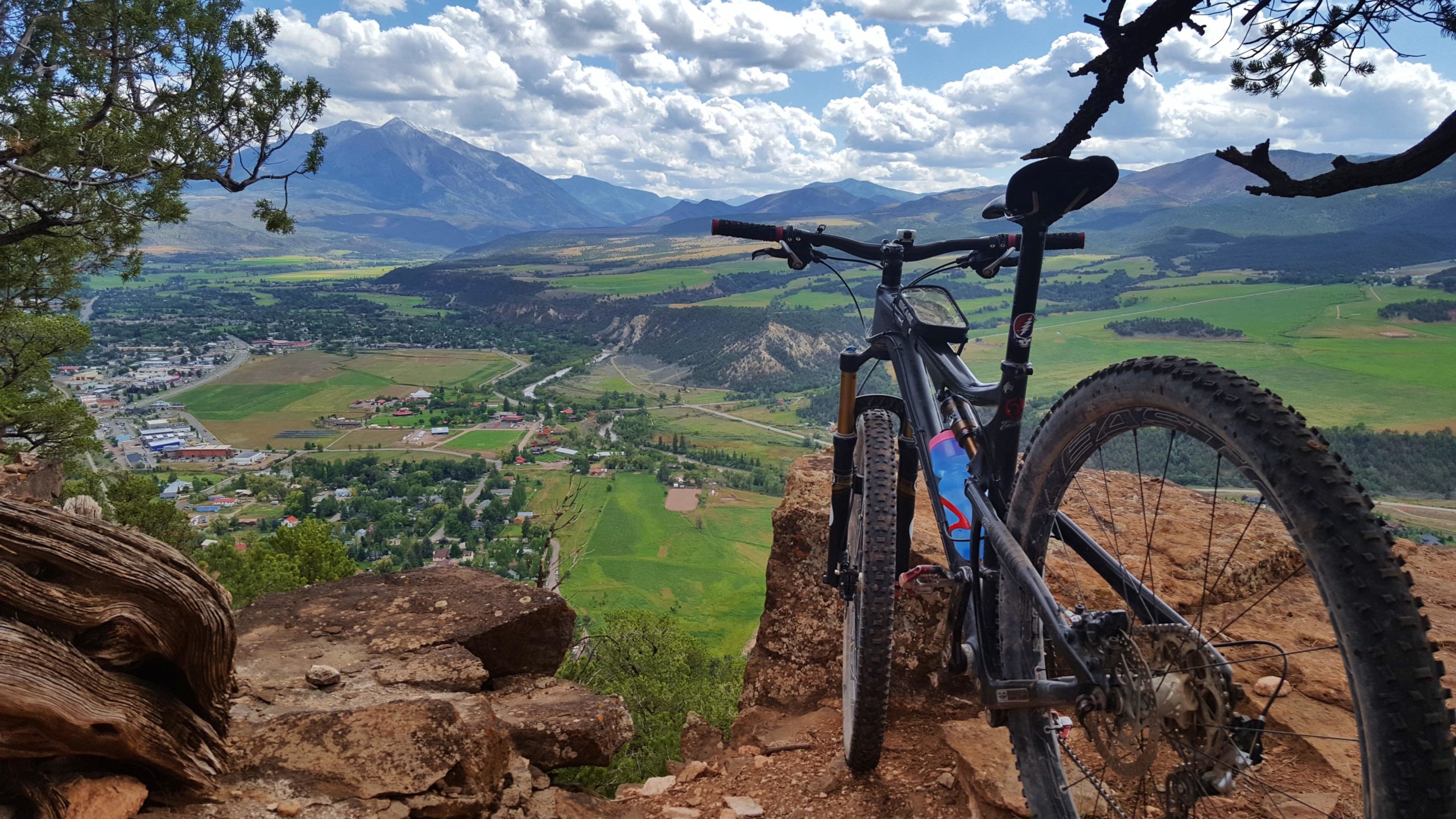 A mountain bike positioned on a rocky ledge overlooking a lush valley filled with fields and a small town. The scene features distant mountains under a partly cloudy sky, showcasing the natural beauty of the landscape. Red Hill mountain bike trail.