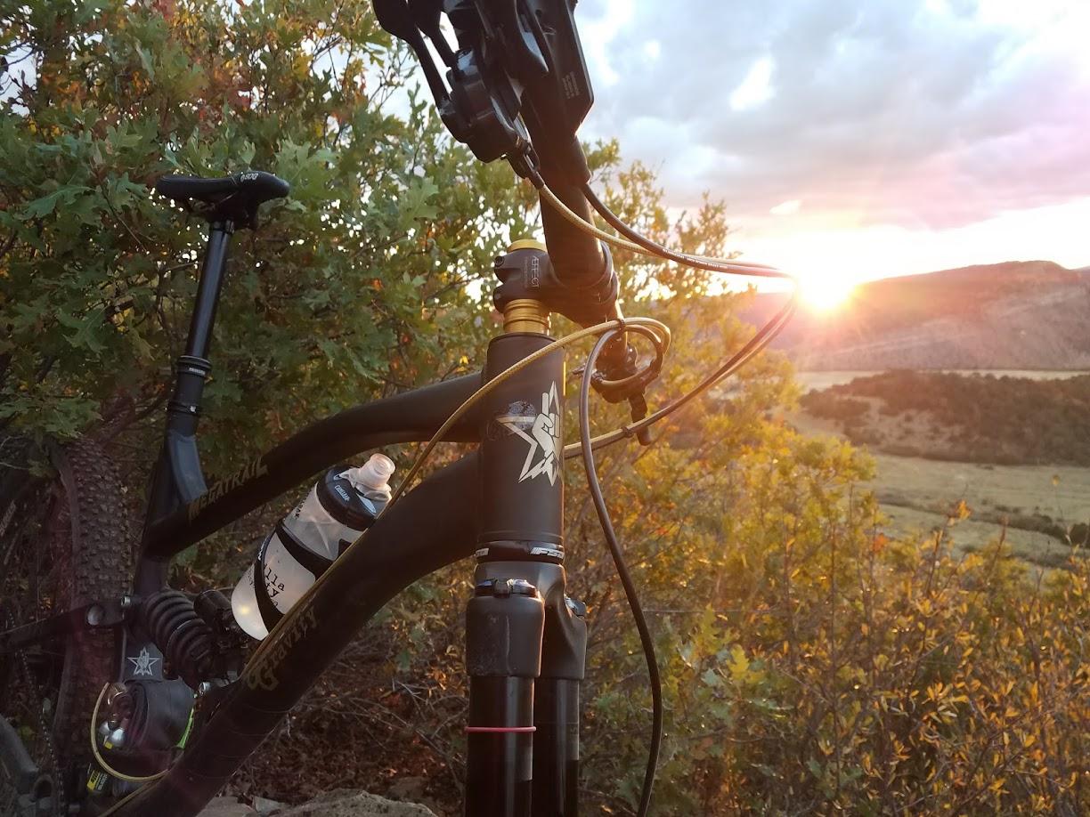Guerrilla Gravity Megatrail: A mountain bike parked near a trail, with a water bottle attached, in the foreground. The sun is setting in the background, casting warm light over the landscape filled with shrubs and hills.