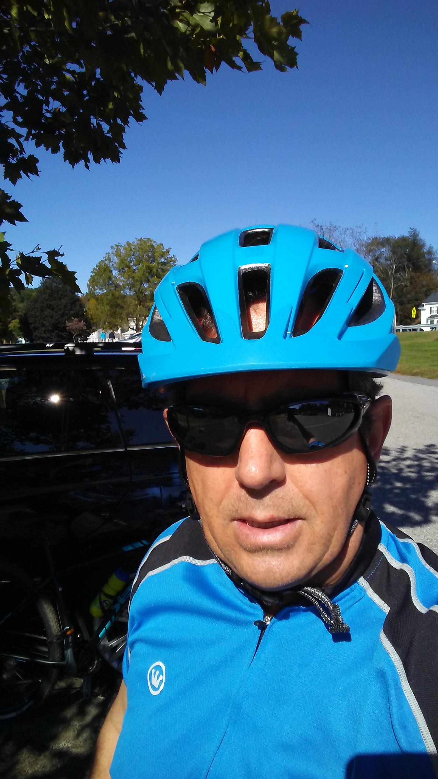 A middle-aged man wearing a blue cycling helmet and sunglasses, looking directly at the camera. He is dressed in a blue and black cycling jersey. In the background, there is a black vehicle and clear blue sky with a few trees visible.