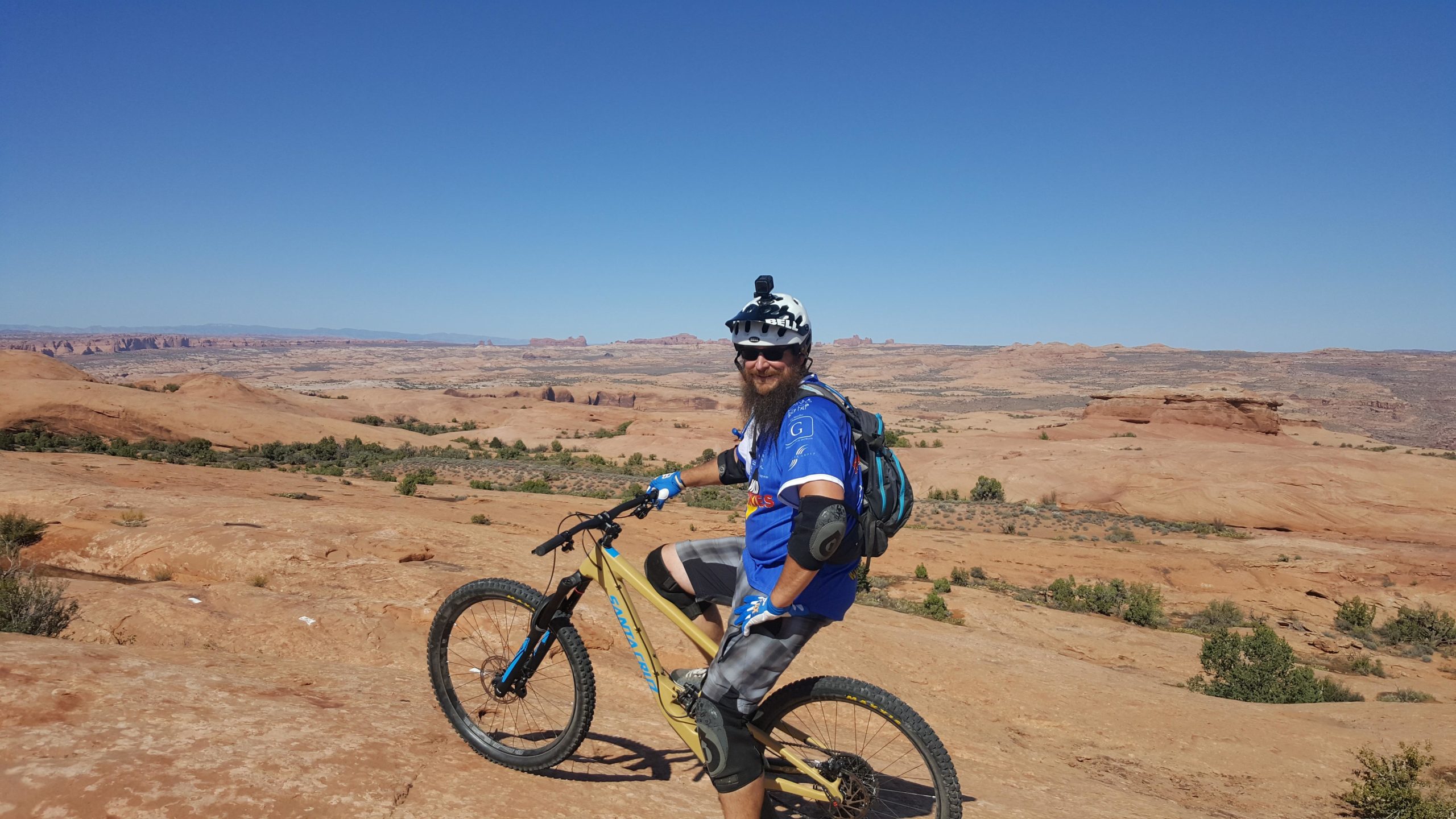 A person with a beard, wearing a blue cycling jersey, helmet, and protective gear, poses on a mountain bike on rocky terrain. They are smiling and looking back at the camera, with a expansive desert landscape and a clear blue sky in the background. Slickrock mountain bike trail.