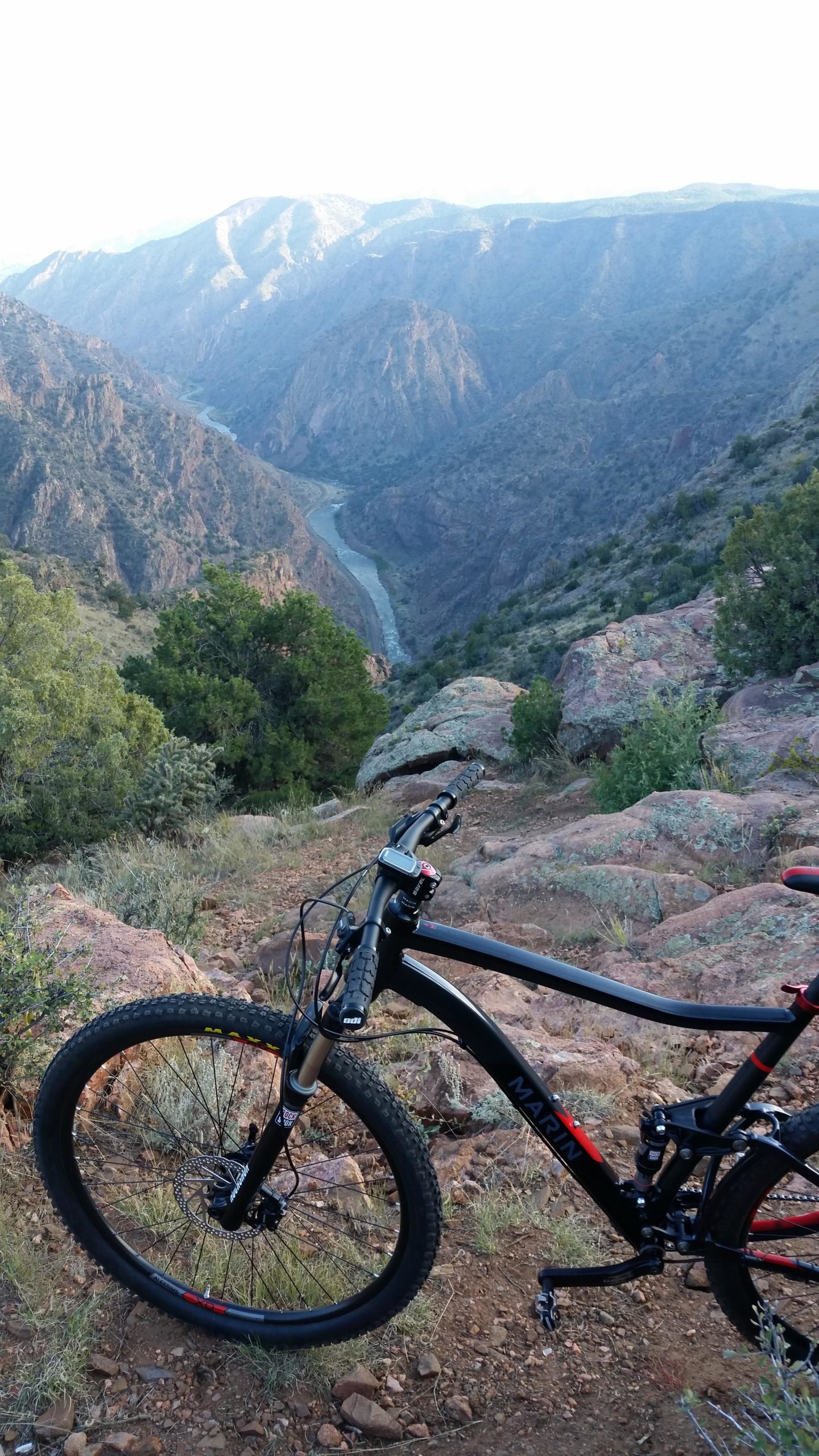 A mountain bike resting on rocky terrain, overlooking a winding river and scenic mountains in the background. The landscape features lush greenery and rugged slopes, suggesting a remote outdoor location ideal for biking and nature exploration. Royal Gorge Park Trail System mountain bike trail.