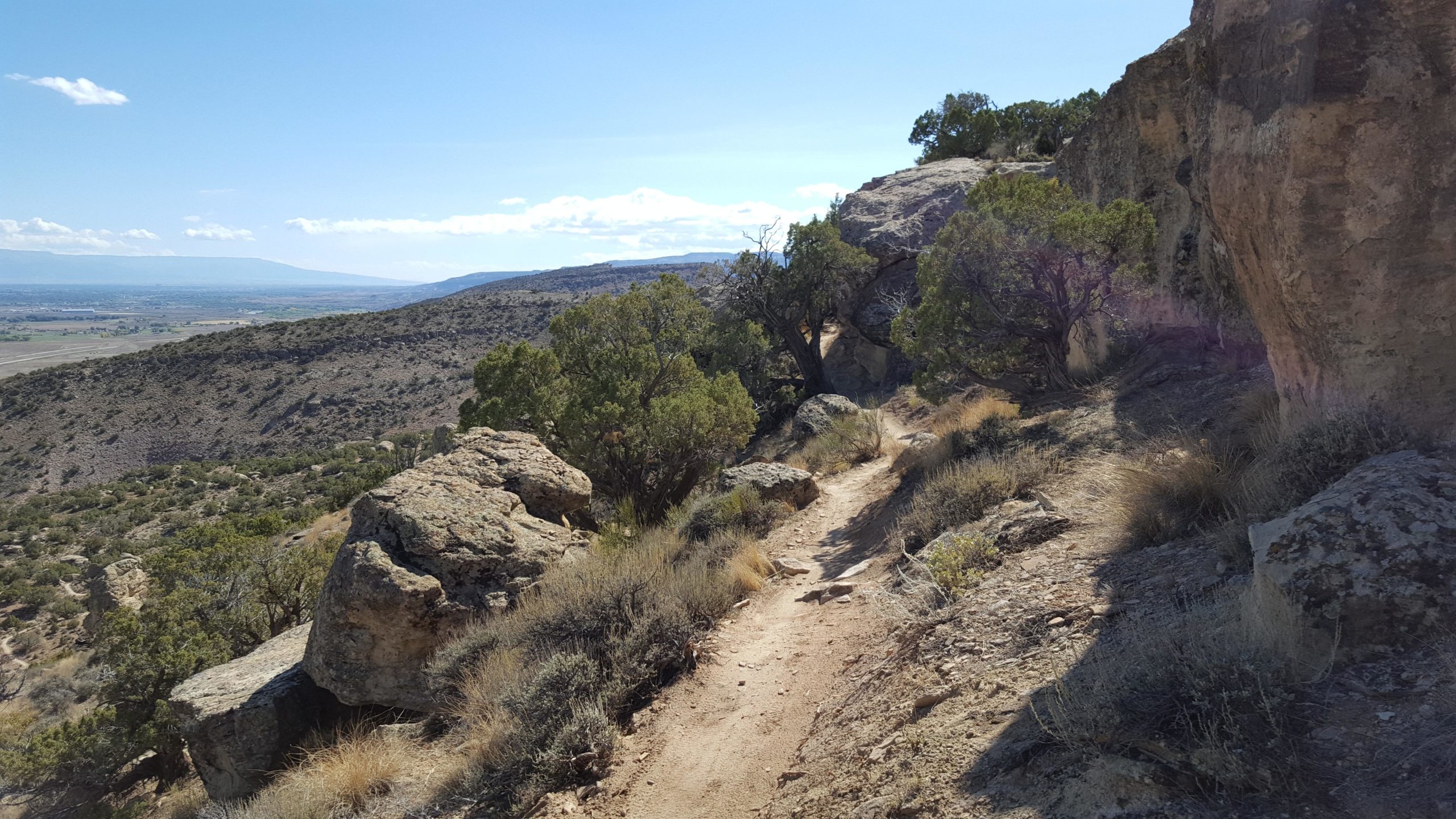 A scenic hiking trail winding through rocky terrain, surrounded by sparse vegetation and trees, with a view of distant hills and a clear blue sky. Hawkeye Trail mountain bike trail.