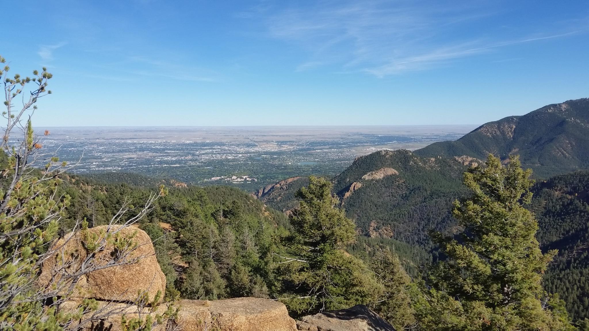 A panoramic view of a mountainous landscape, featuring lush green forests and rocky outcrops, with a vast valley visible in the distance under a clear blue sky. Stratton Open Space / The Chutes mountain bike trail.