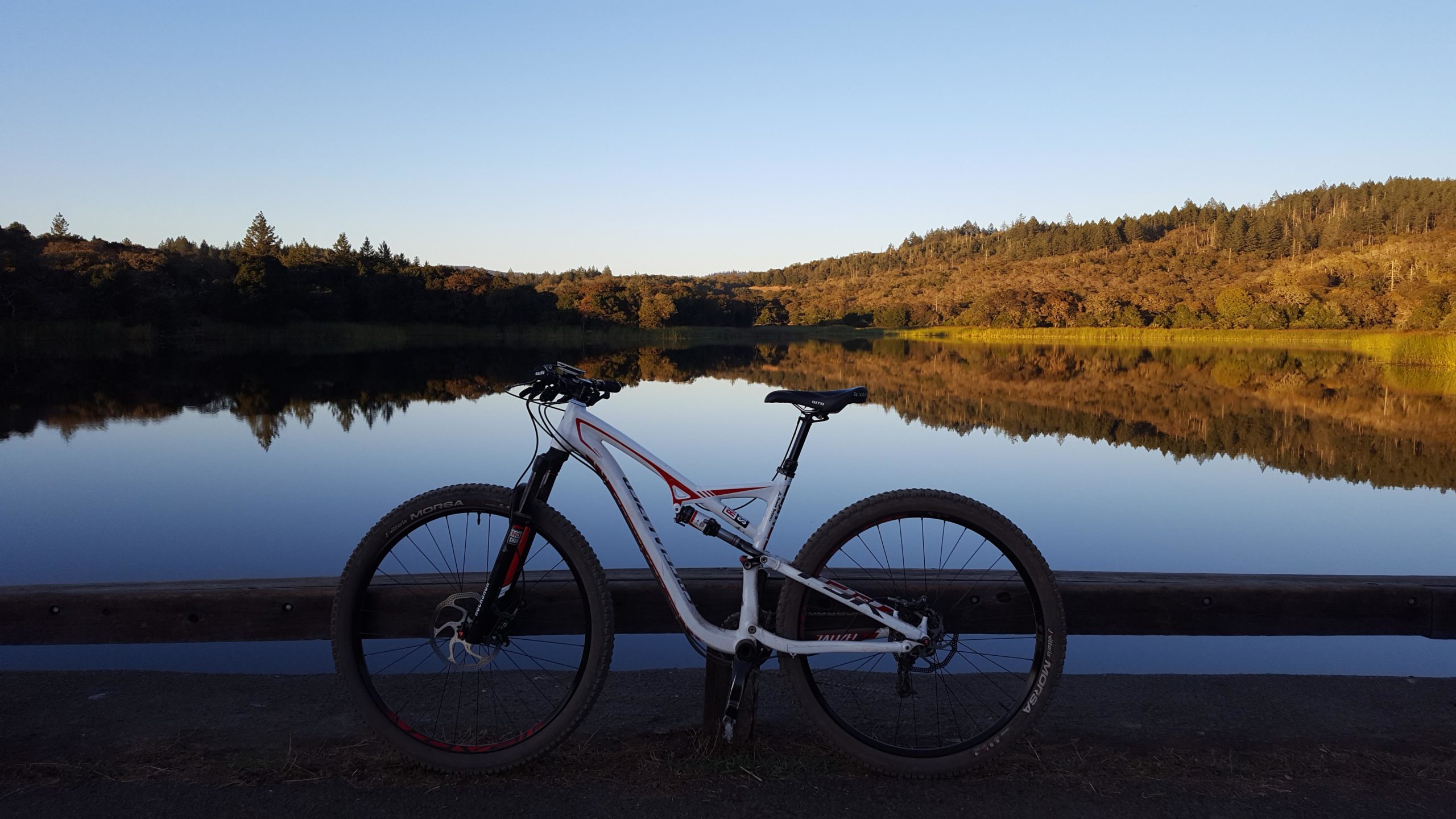 A mountain bike parked on a wooden railing by a calm lake, surrounded by trees reflecting in the water. The scene is bathed in soft evening light under a clear blue sky. Annadel State Park mountain bike trail.