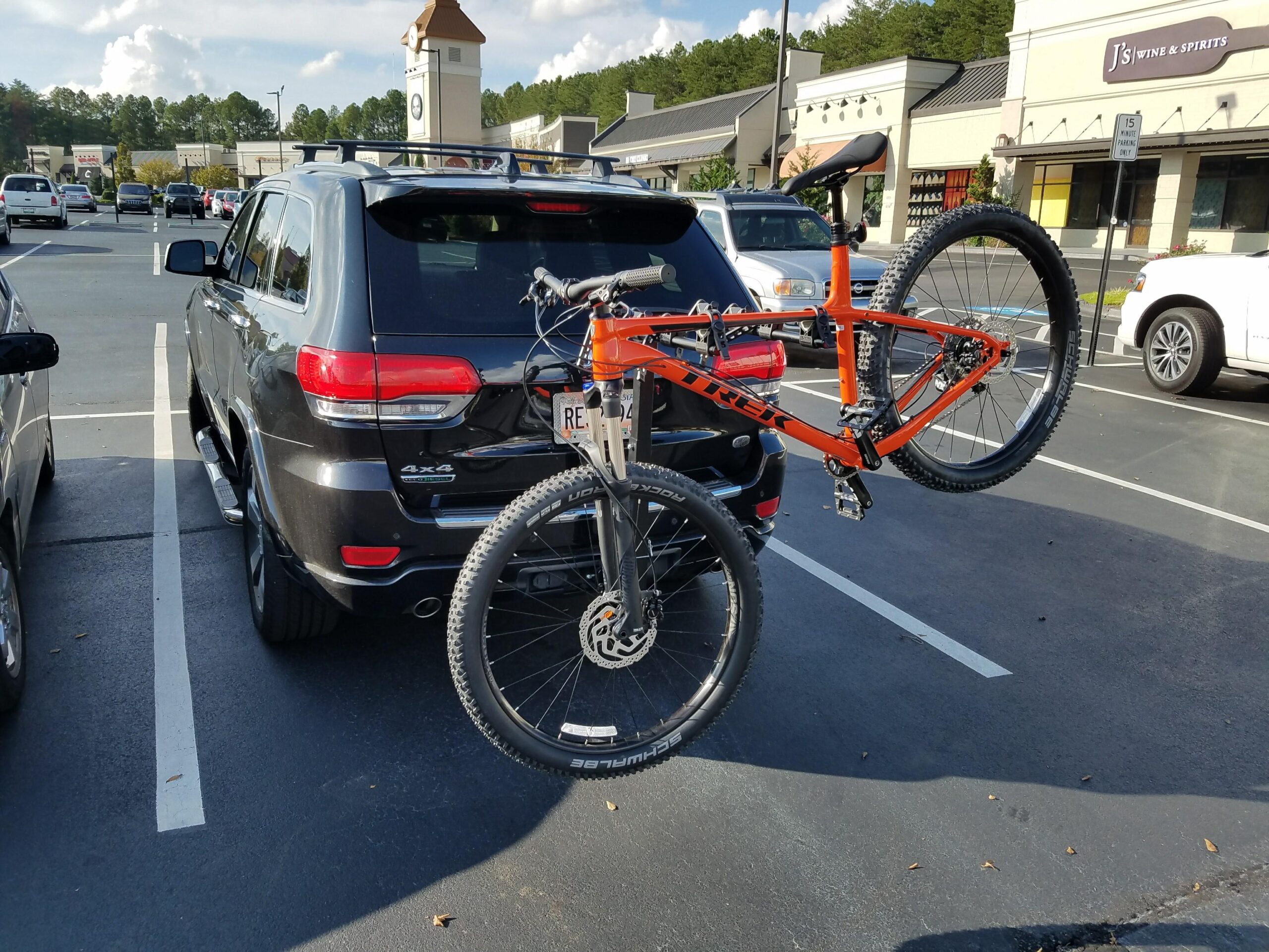 Trek Roscoe 8: A black SUV parked in a shopping area parking lot, featuring a mounted orange mountain bike on a bike rack at the rear. In the background, several other vehicles and retail buildings are visible under a partly cloudy sky.