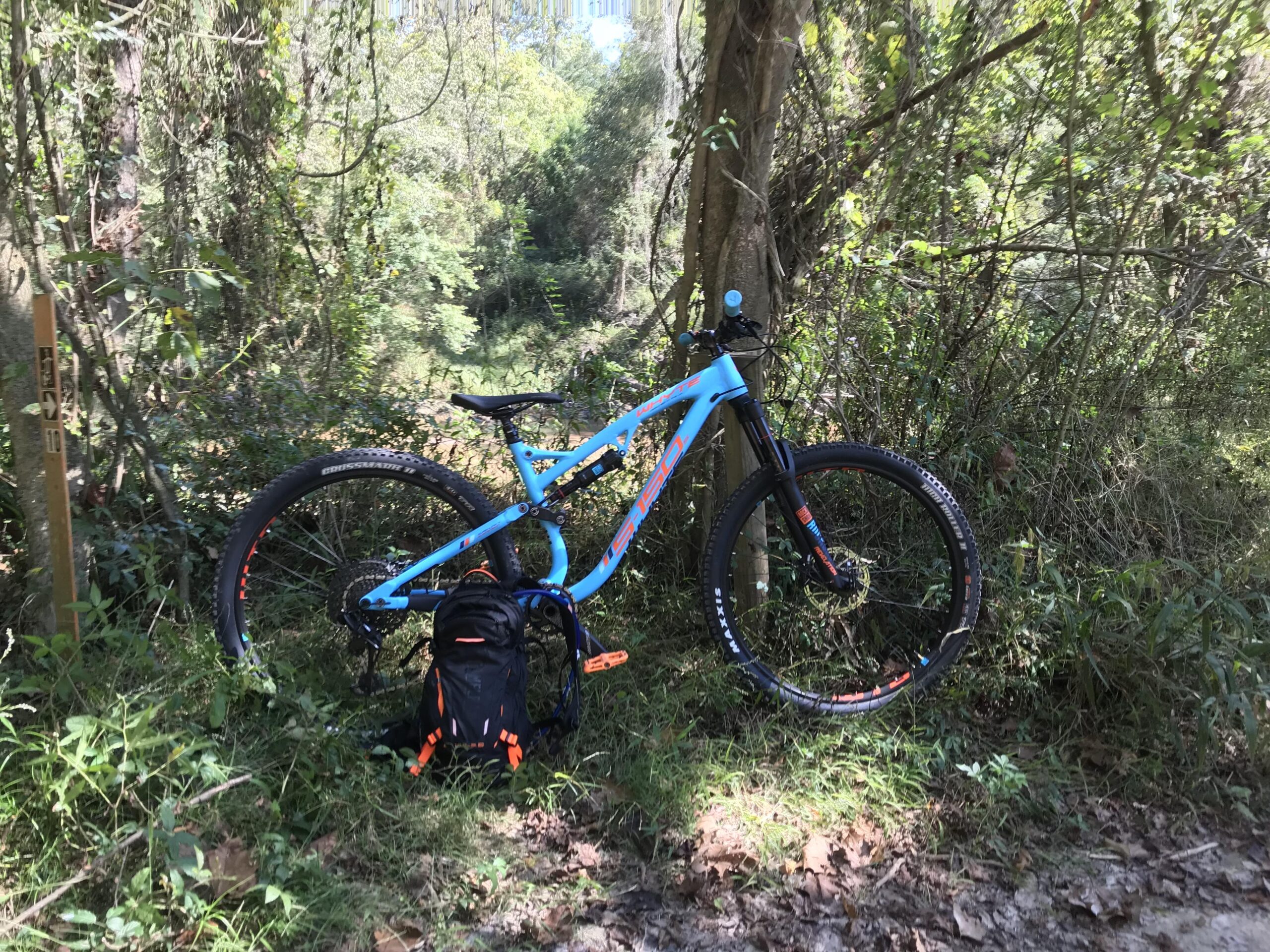 Whyte S-150s: A blue mountain bike with orange accents rests on the forest floor next to a black backpack, surrounded by dense greenery and trees. A trail marker is visible in the background.