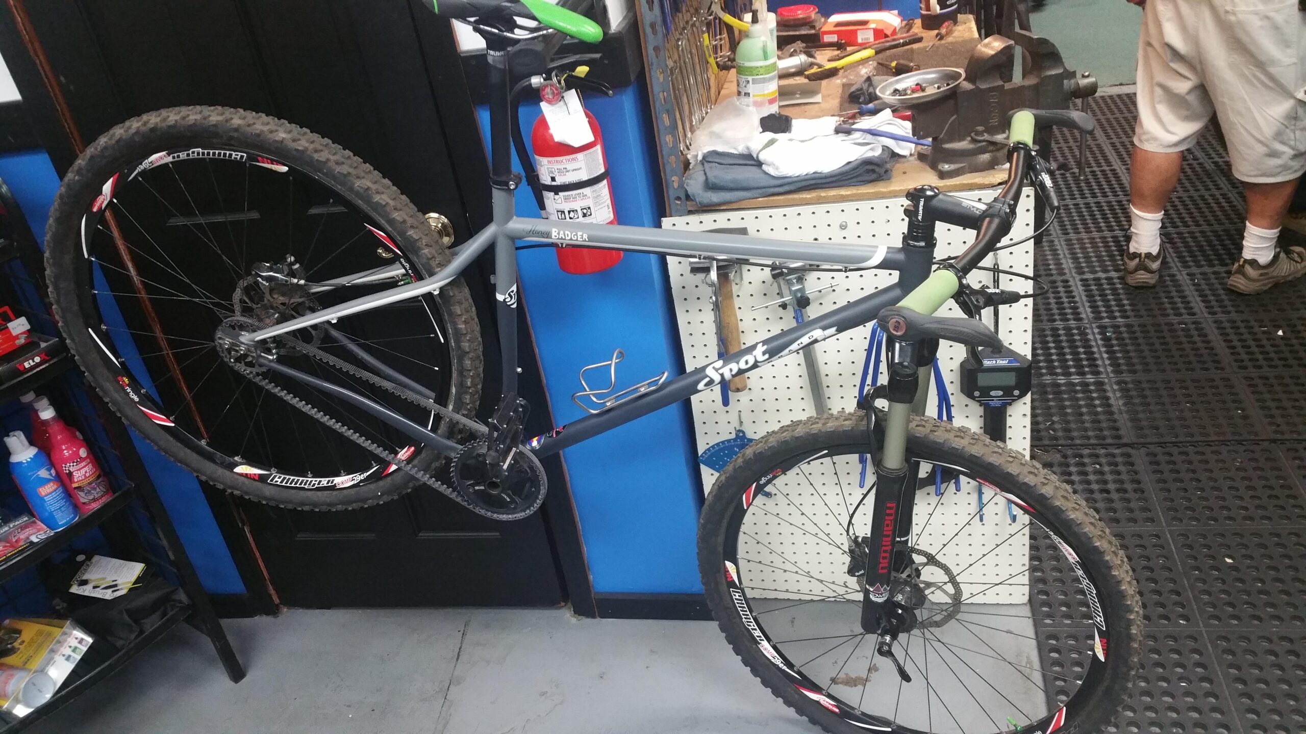 SPOT Honey Badger Singlespeed: A mountain bike leaning against a workbench in a bike shop. The bike features a gray frame with green handlebar grips and is equipped with knobby tires showing some dirt. Behind it, there are shelves with various bike maintenance supplies, including bottles and tools, while a person is partially visible in the background.