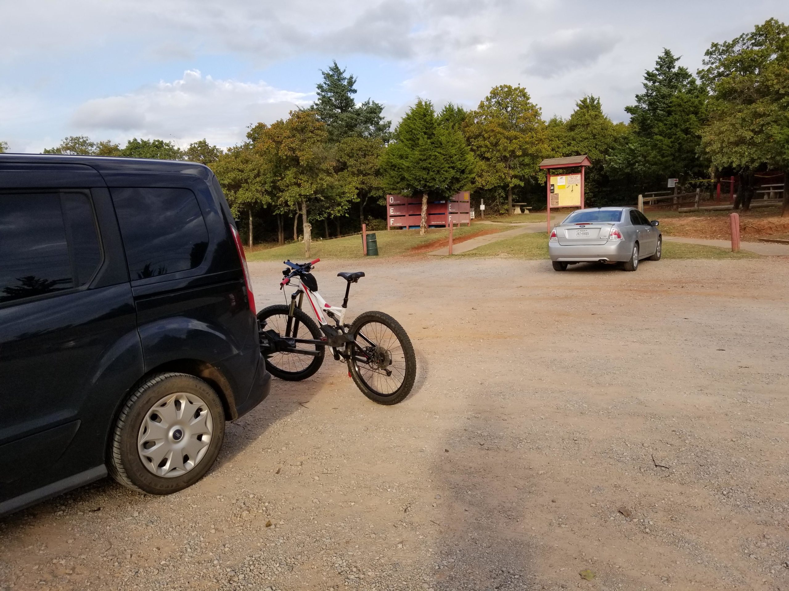 A black van and a white mountain bike are parked in a gravel lot surrounded by trees. In the background, a silver car is parked, and a wooden information board is visible, indicating a recreational area. The scene is set under a cloudy sky with a few patches of blue visible. Lake Stanley Draper mountain bike trail.